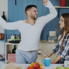 Attractive young joyful couple have fun dancing and singing while set the table for breakfast in the kitchen at home